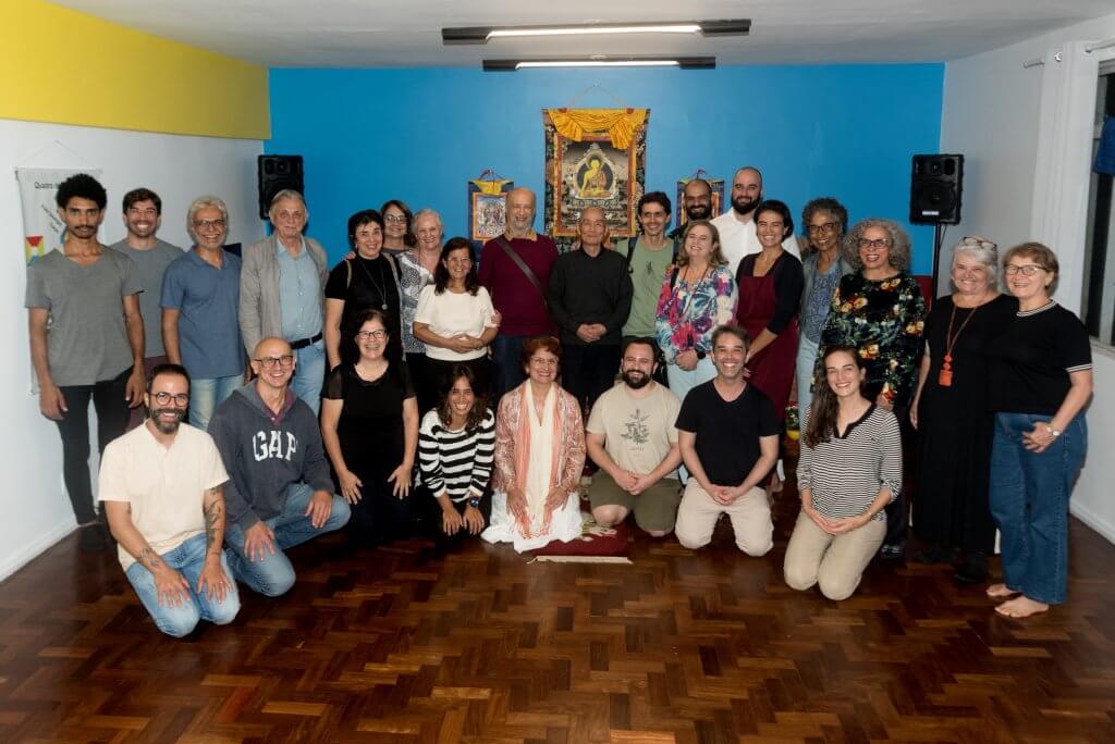 Mestre Tokuda está realizando uma prostração diante do altar na sala do CEBB BH. Um homem participante do encontro pode ser visto na foto com as mãos em prece, à esquerda. Alguns objetos compõem a cena, como uma televisão lateral, o zafu utilizado para meditar, um jarro de flores e cadeiras. No altar há fotos de mestres e uma estátua dourada do Buda Shakyamuni em posição de lótus no centro do altar. 