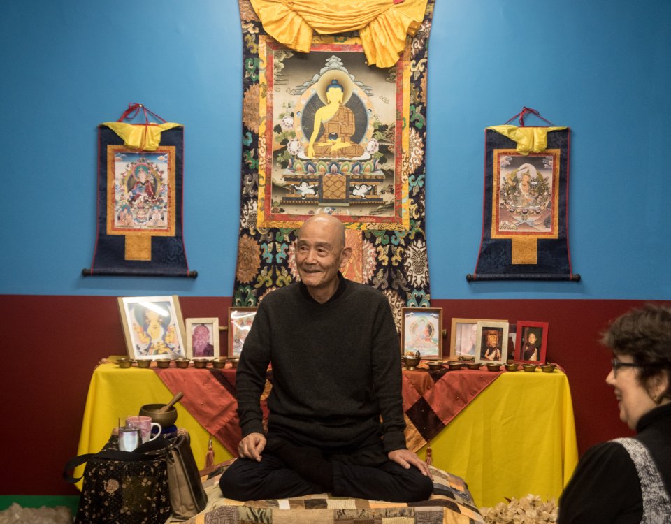 Mestre Tokuda está sentado ao centro da foto em posição de lótus, radiante e sorridente, com olhar compassivo. Atrás dele há um altar com fotos de vários mestres, além de imagens de Budas. À esquerda observamos alguns objetos, como sino e xícara de chá. À direita uma mulher participante do evento aparece sorrindo.